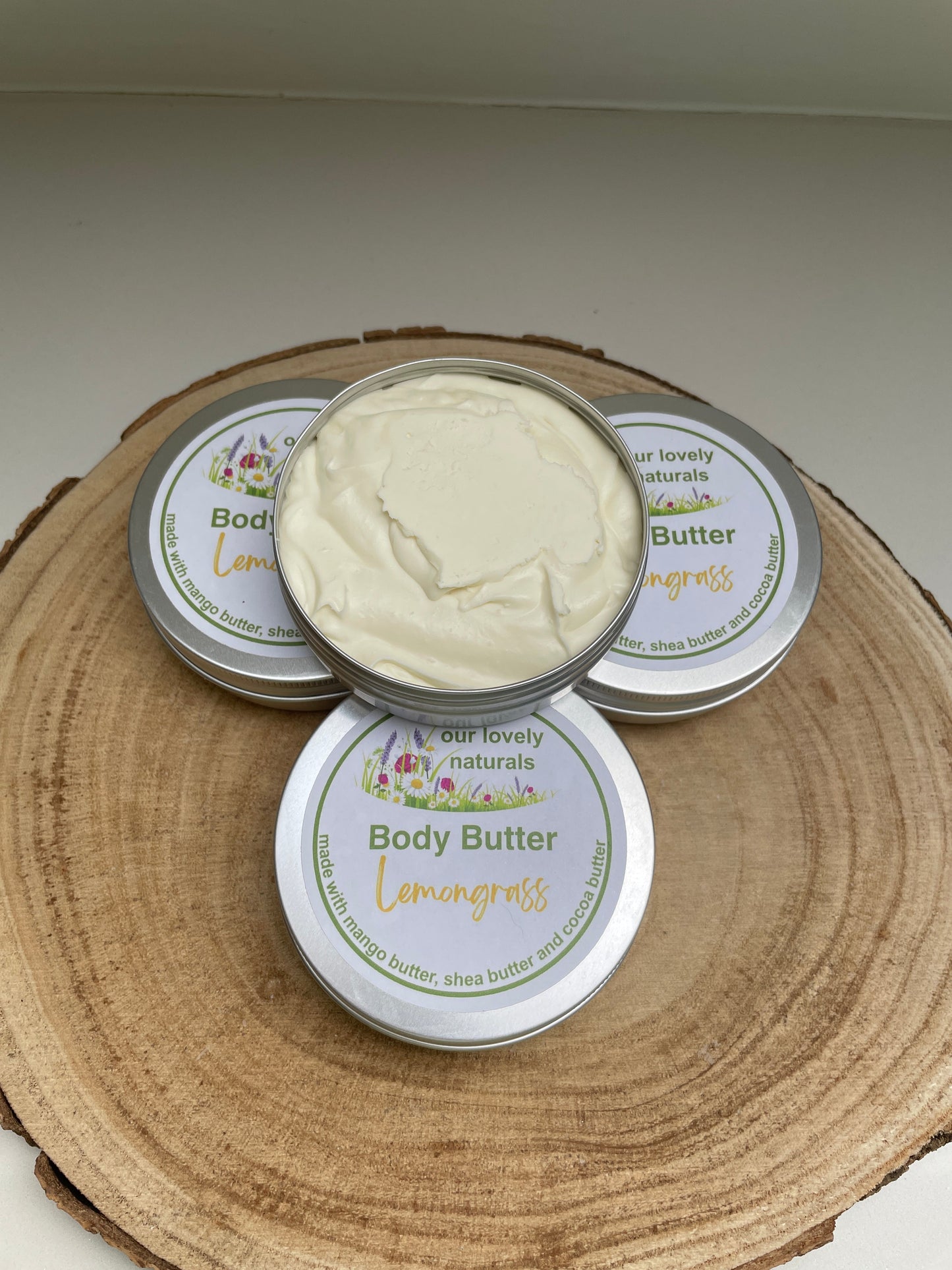 Four tins of 100% natural, handmade Lemongrass body butter, one with the lid off so that the viewer can see the content.  All on a wooden board.