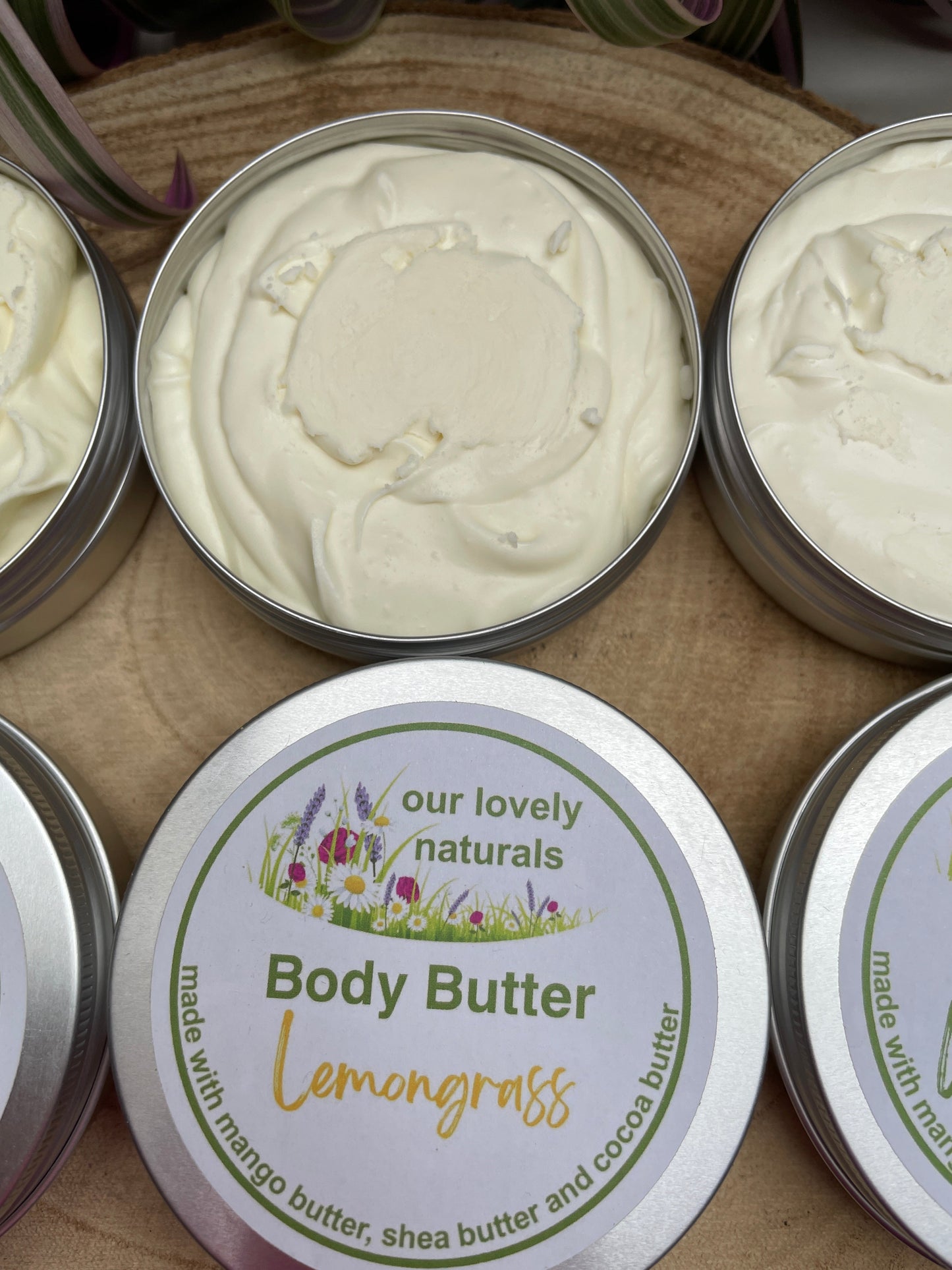 Two tins of 100% natural, handmade Lemongrass body butter, one with the lid off so that the viewer can see the content.  All on a wooden board.