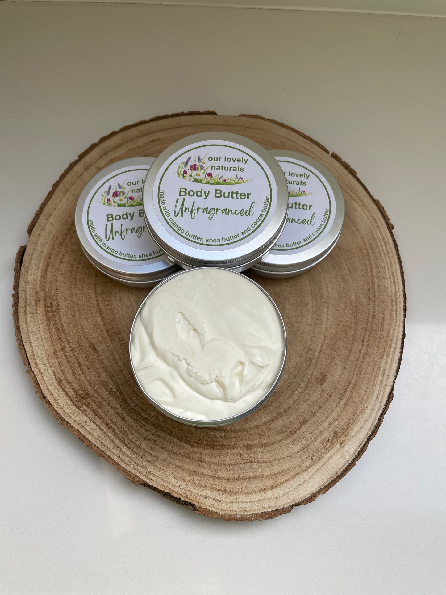 Four tins of 100% natural, handmade unfragranced body butter, one with the lid off so the viewer can see the content,  on a wooden board.